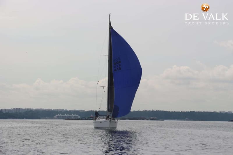 Een witte zeilboot met een blauw zeil vaart op het water, met een bebouwde kustlijn op de achtergrond. — Hanse 415