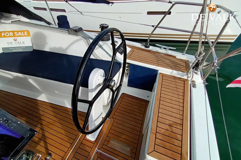 Een boot met een stuurwiel, teakhouten dek en blauwe kussens, met een "For Sale" bord van De Valk Yacht Brokers. — Hanse 415
