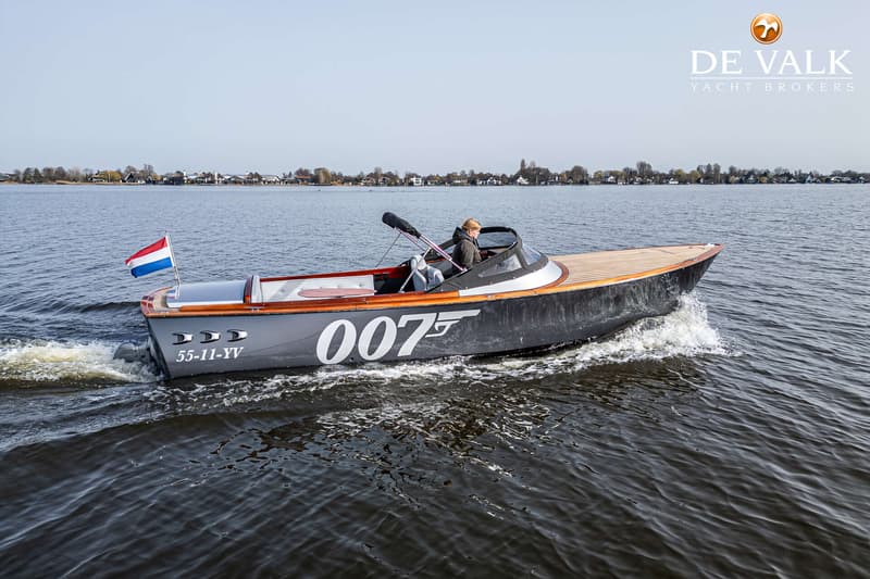 Een grijze motorboot met houten dek en de opdruk "007" vaart op een meer met een Nederlandse vlag aan de achtersteven.
