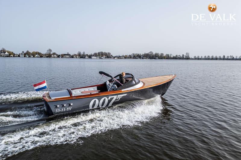 Een donkere boot met houten dek en de cijfers "007" op de zijkant vaart over water, met een persoon aan het roer.