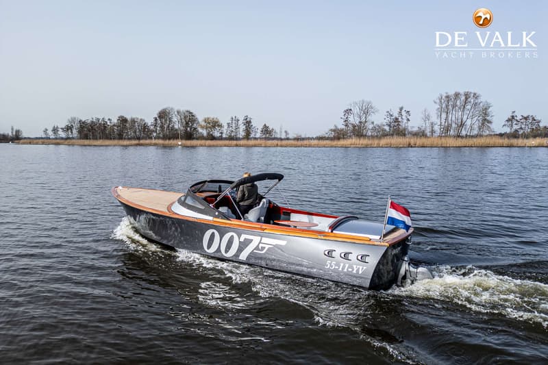 Een grijze boot met "007" en "55-11-YV" op de zijkant vaart op het water met een Nederlandse vlag aan de achterkant.