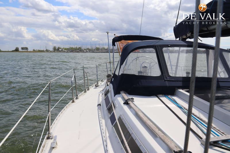 Een witte zeilboot met een donkerblauwe buiskap vaart op water met een jachthaven op de achtergrond. — Beneteau First 405
