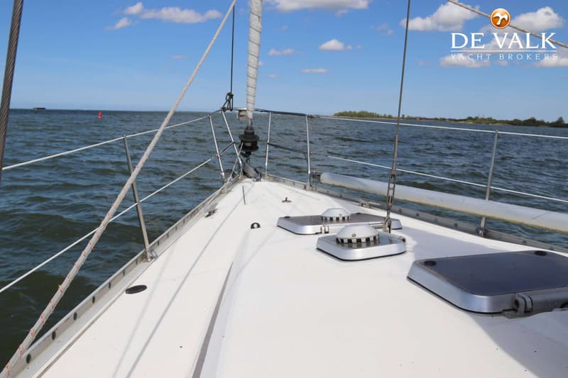De boeg van een zeilboot met een opgerold zeil, reling en luiken, varend op water met een eiland op de achtergrond.