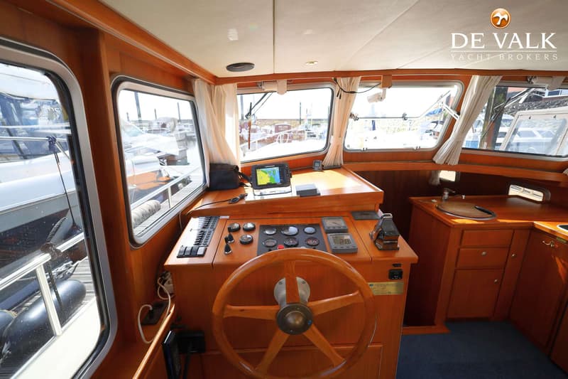 De foto toont het interieur van een boot met een houten stuurwiel, navigatieapparatuur en een kleine kombuis.