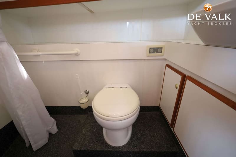 Een witte toiletpot met een beige zitting staat op een donkere vloer, met een toiletborstel ernaast en een douchegordijn…