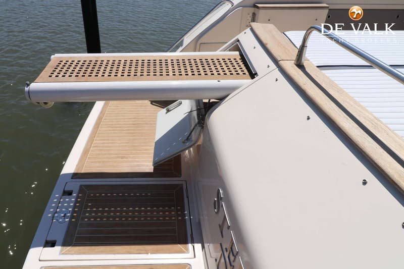 Een deel van een boot met een teakhouten dek, een uitschuifbaar platform en een reling, met water op de achtergrond.