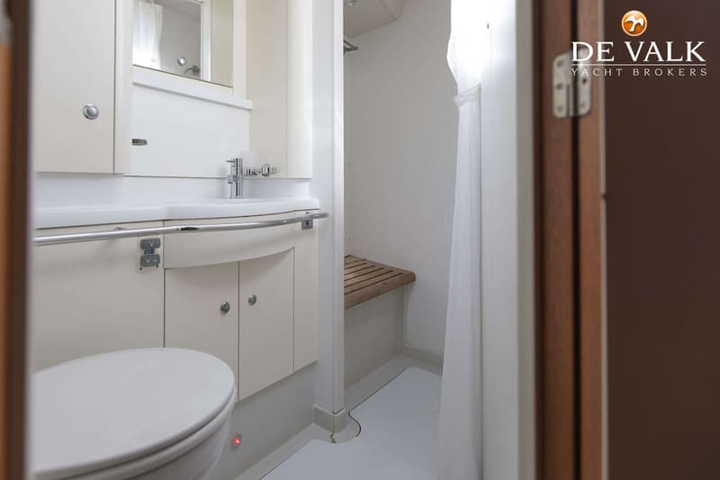 Een witte badkamer met toilet, wastafel, spiegel en een douchecabine met houten bankje en wit douchegordijn.