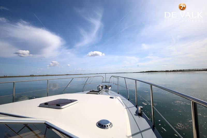 De foto toont de boeg van een witte boot met een roestvrijstalen reling, varend op rustig water onder een blauwe lucht.