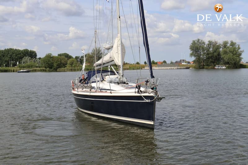 Een donkerblauwe zeilboot met witte strepen vaart op een waterweg met een man aan het roer. — Dehler 39 SQ