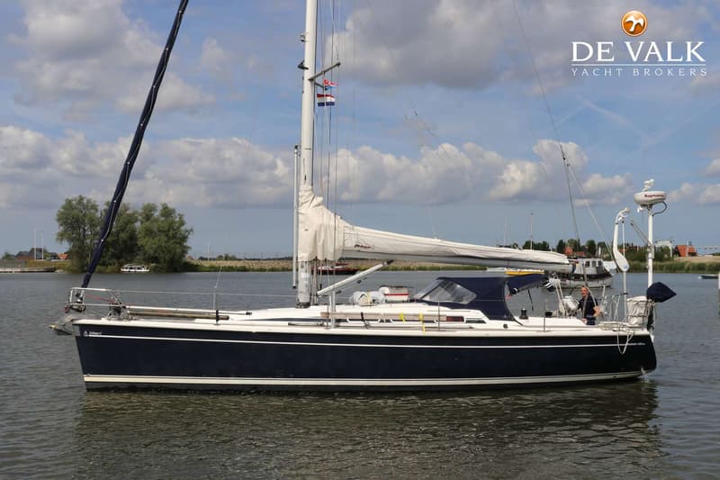 Een donkerblauwe zeilboot met witte strepen vaart op het water, met een man aan het roer en een zeil met "Dehler" erop.