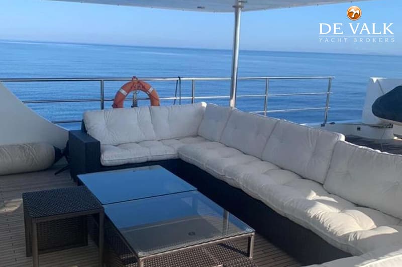 Een hoekbank met witte kussens en twee salontafels op een houten dek met de zee op de achtergrond.