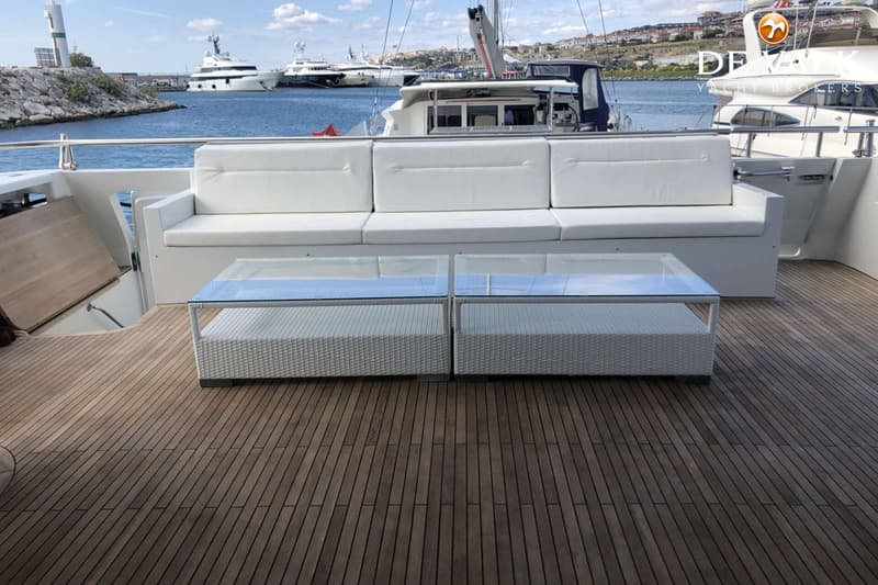 Een teakhouten dek met een witte bank en twee witte salontafels met glazen blad, met op de achtergrond een haven.