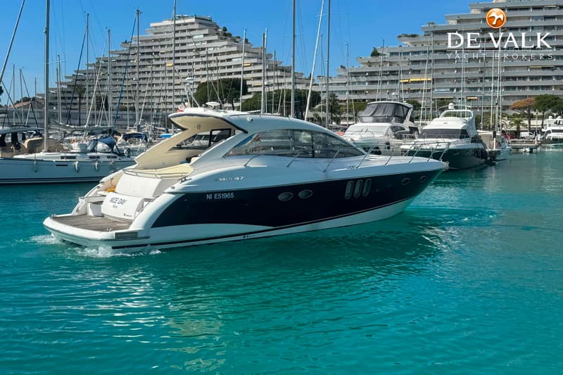 Een witte motorboot met een donkerblauwe romp vaart in een haven met helderblauw water, omringd door andere boten en…