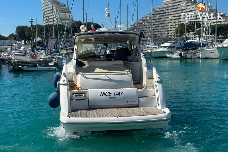 Een witte motorboot met de naam "NICE DAY Nice" op de spiegel vaart in een haven met gebouwen op de achtergrond.