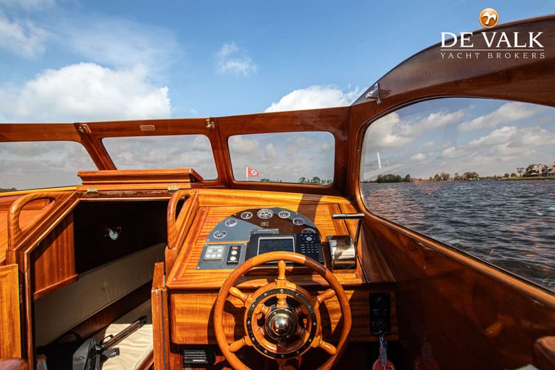 Een houten stuurhut met een stuurwiel, navigatie-instrumenten en uitzicht op water en landschap.
