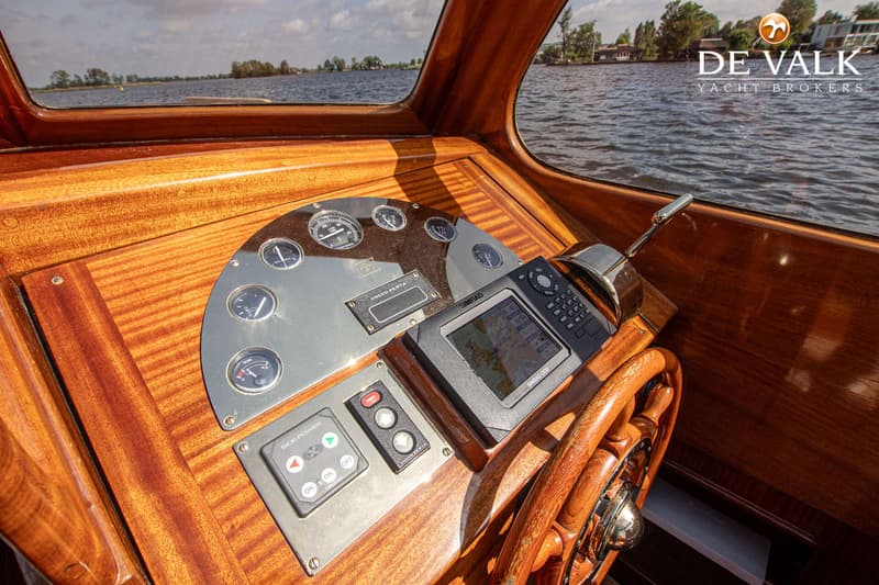Een houten dashboard met diverse meters, een Simrad plotter, bedieningspanelen en een stuurwiel, met water en landschap op…