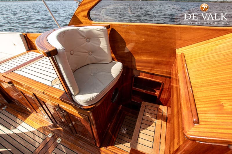 Een houten bootinterieur met een beige kussen op een zitje, een trapje naar beneden en een logo van "DE VALK YACHT BROKERS".