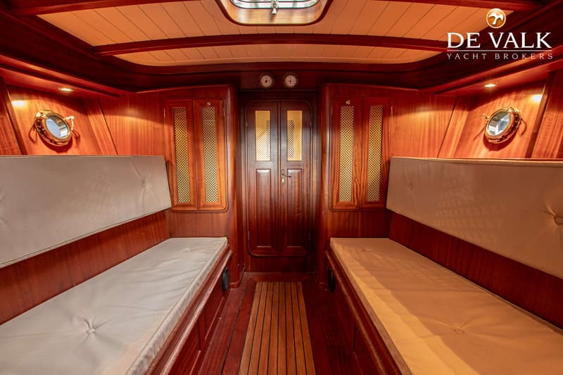 De foto toont het interieur van een boot met houten wanden, twee banken met lichte kussens en een deur aan het einde.