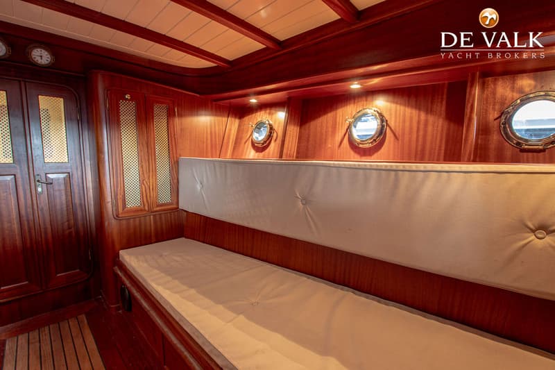 Een houten interieur met een bank, patrijspoorten en kasten, met het logo van "DE VALK YACHT BROKERS" rechtsboven.