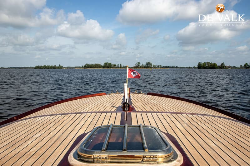 De foto toont de boeg van een houten boot met een rood vlaggetje met een witte 'B' erop, varend op een meer.