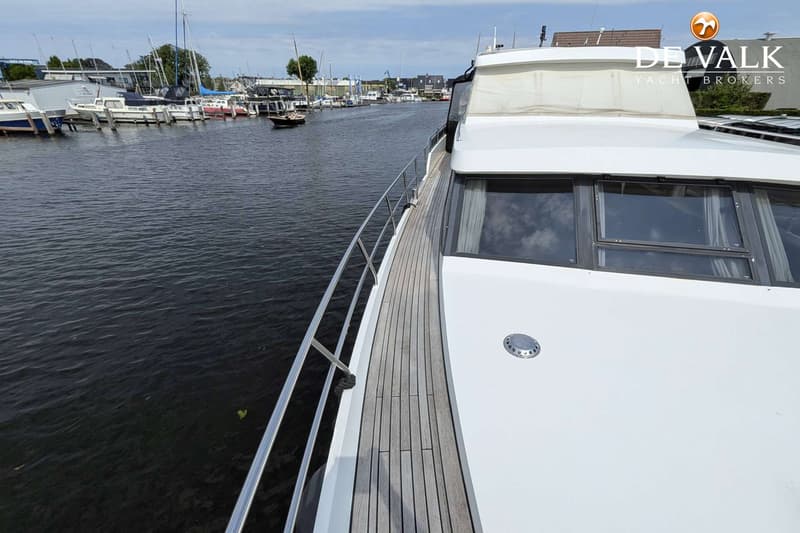 Een witte boot met een teakhouten dek en een zilveren reling vaart op een kanaal langs andere boten.