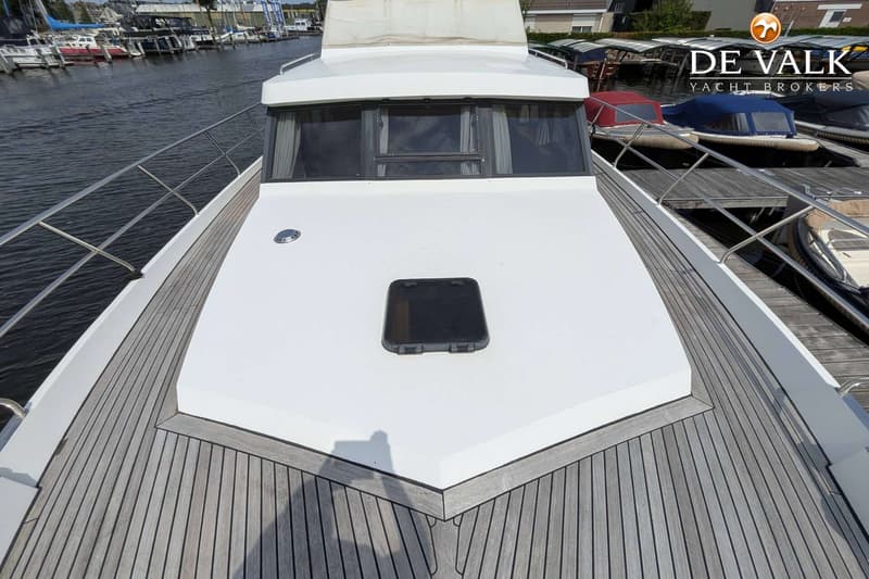 Een witte boot met een houten dek en een zwart luik op de boeg, liggend aan een kade met andere boten.
