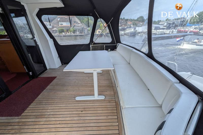 Een dek van een boot met een witte tafel, een witte bank en een uitzicht op water en huizen. — Van der Heijden Elegance 1500