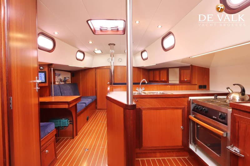 De foto toont het interieur van een boot met een houten vloer, een kombuis met fornuis en spoelbak, en een zithoek.
