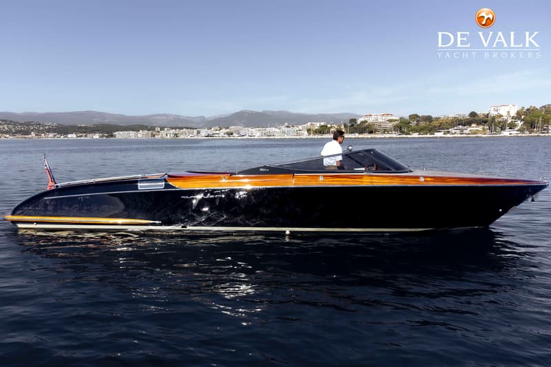 Een donkerblauwe speedboot met houten accenten en een persoon aan het roer vaart op rustig water, met een kustlijn op de…