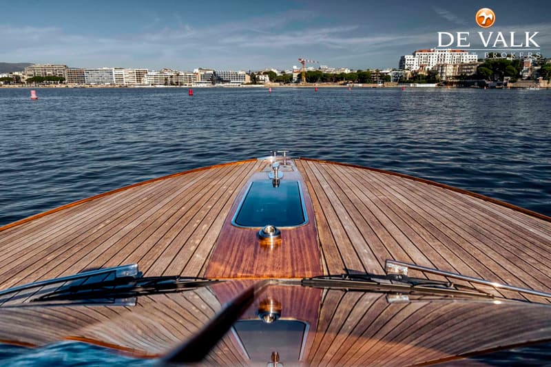 Een houten dek van een boot met een patroon van lange planken, een raam en een metalen bolder, varend op water met een stad…
