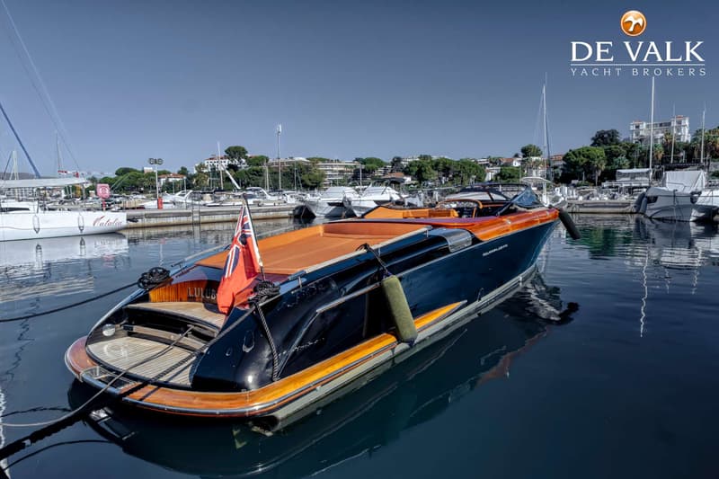 Een donkerblauwe speedboot met houten accenten en een oranje interieur ligt afgemeerd in een jachthaven.