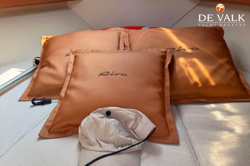 Drie bruine kussens met het woord "Riva" erop, een beige pet en een oranje reddingsboei zijn zichtbaar.