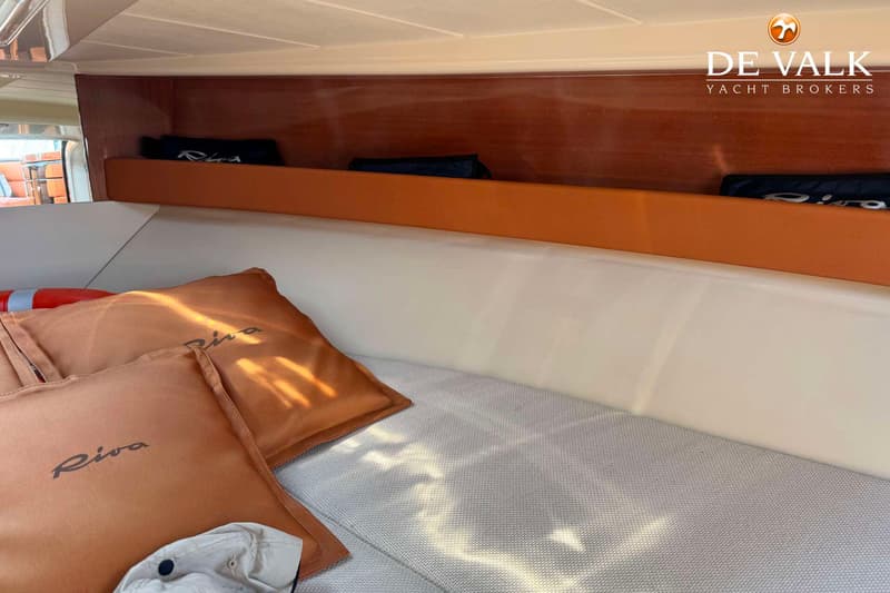 Een interieur van een boot met een bed, kussens met het woord "Riva" erop, en een houten wand met een plank.