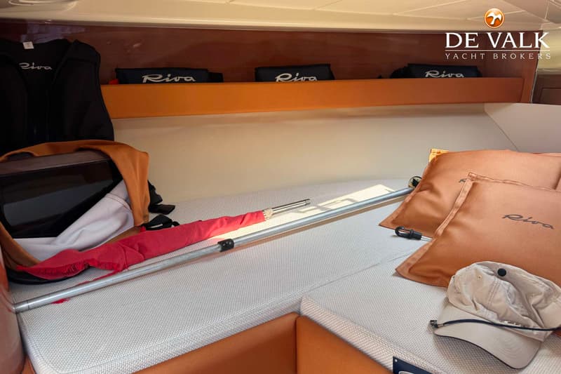 De foto toont een interieur van een boot met een bed, kussens, een pet, een stok en opbergvakken met Riva-logo's.
