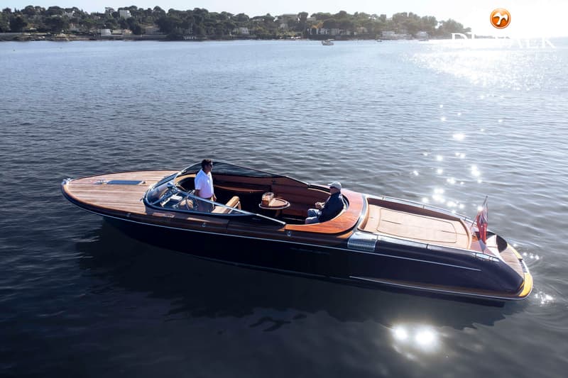 Een donkerblauwe motorboot met houten dek en interieur vaart op kalm water met twee mannen aan boord. — Riva Aquariva Cento