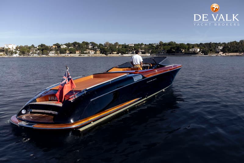 Een donkerblauwe motorboot met houten accenten en een man aan het roer vaart op rustig water, met een kustlijn op de…