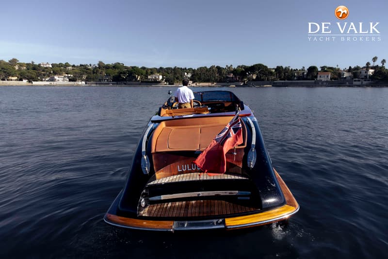 Een man bestuurt een donkere boot met bruine bekleding en een Britse vlag aan de achterkant, varend op rustig water.