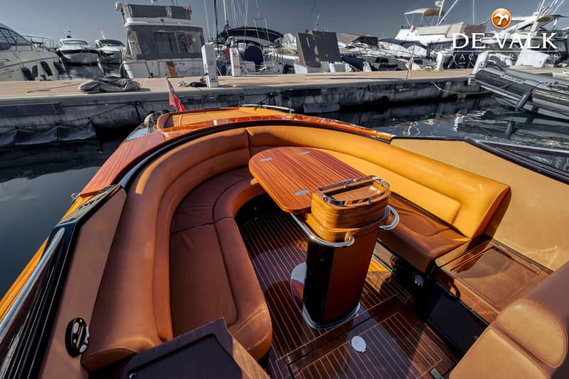 Een bovenaanzicht van een boot met een houten dek, bruine leren banken en een houten tafel, liggend in een haven.