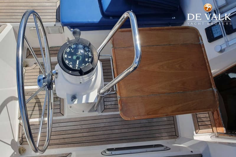 Een stuurwiel, kompas, houten tafel en een blauw kussen zijn zichtbaar op een teakhouten dek.