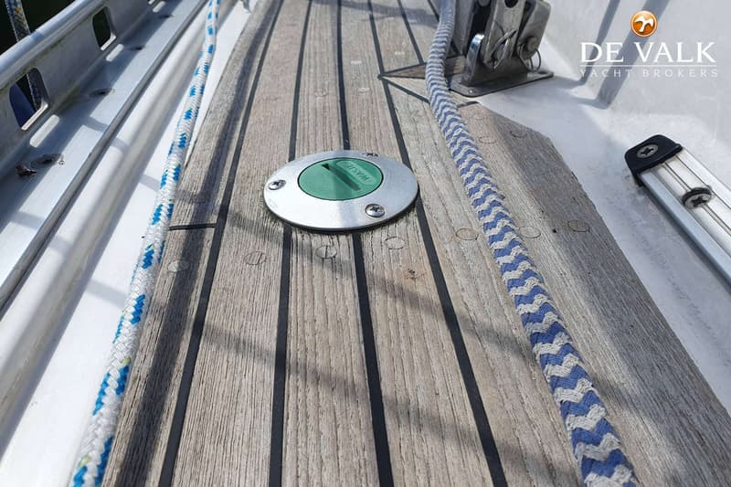Een close-up van een teakhouten dek met een groen "WASTE" deksel en een blauw-wit touw.