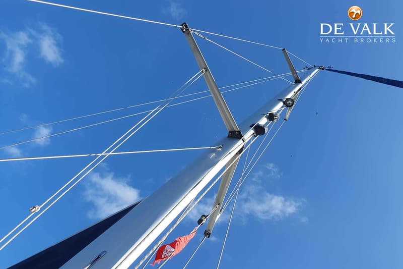 Een mast van een zeilboot met een opgerold zeil en een rode vlag, gezien tegen een blauwe lucht met enkele wolken.