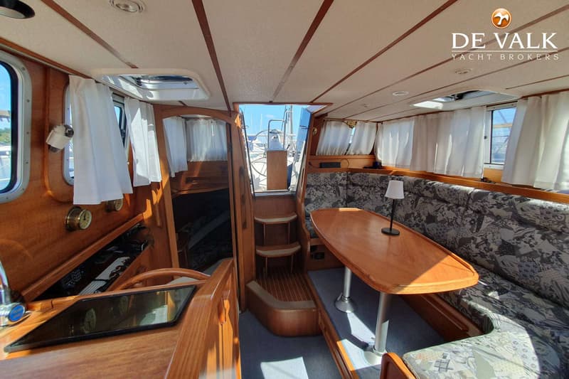 Het interieur van een boot met houten afwerking, een tafel, banken met patroon en een stuurwiel zichtbaar door een opening.