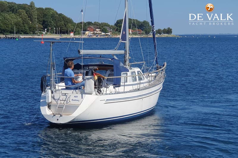 Een witte zeilboot met blauwe strepen vaart op blauw water, met een man aan het roer en een kustlijn op de achtergrond.
