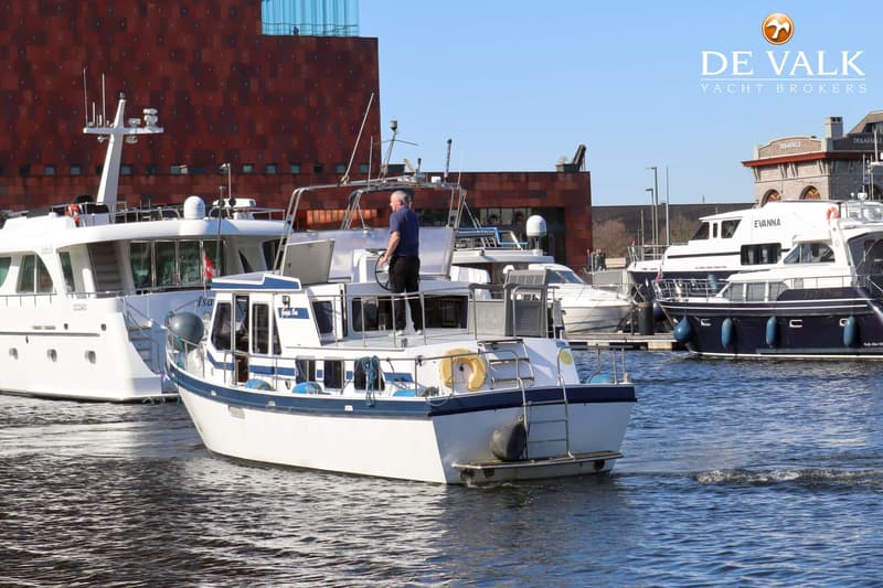 Een man staat aan het roer van een witte motorboot met blauwe accenten, omringd door andere boten in een haven.