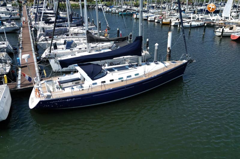 Een donkerblauwe zeilboot met een teakhouten dek ligt aangemeerd in een jachthaven vol met andere zeilboten.