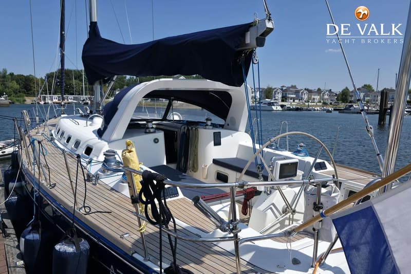 Een zeilboot ligt afgemeerd aan een steiger, met een blauw zeil en een houten dek. — Beneteau First 47.7