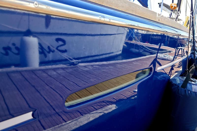 Een detailfoto van de glimmende blauwe romp van een boot met een houten dek en een patrijspoort. — Beneteau First 47.7