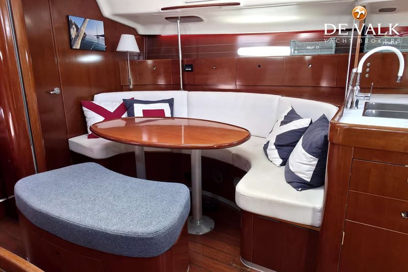 Een houten interieur van een boot met een witte bank, een ovale tafel, kussens en een gootsteen. — Beneteau First 47.7