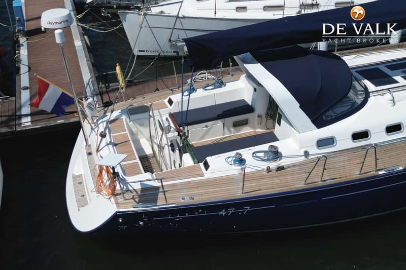 Een blauw-witte zeilboot met teakhouten dek ligt aangemeerd, met een Nederlandse vlag aan de achtersteven.