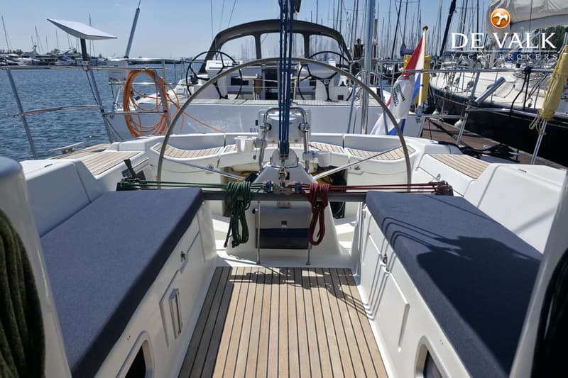 De foto toont de kuip van een zeilboot met teakhouten vloer, blauwe kussens en een stuurwiel, met andere boten op de…
